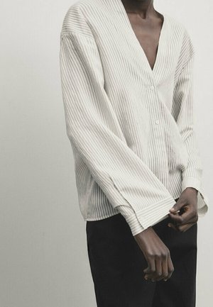 Person wearing a loose, cream-colored, vertically striped buttoned shirt with long sleeves and black pants, adjusting the cuff.