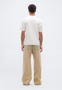 Άνδρας που στέκεται με την πλάτη στην κάμερα, φοράει μια απλή λευκή μπλούζα και χαλαρά μπεζ παντελόνια, με φόντο λευκό.