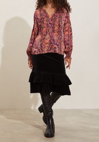 Blusa a maniche lunghe con motivo floreale nei toni di rosa, arancione e viola, con scollatura a V. Abbinata a una gonna in velluto nero con orlo arricciato e stivali al ginocchio.
