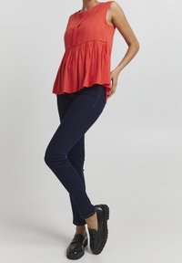 Top sin mangas rojo con un patrón texturizado y un dobladillo fruncido, combinado con jeans ajustados oscuros y mocasines negros. Un atuendo simple y casual.