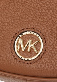 Brun texturerad läderväska med en guldcirkulär logotyp där "MICHAEL KORS" omger bokstäverna "MK". Släta accenter och sömmar synliga.