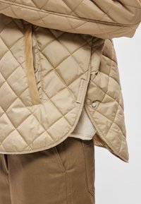 Μπεζ καπιτονέ μπουφάν με διαμάντι σχέδιο, διαθέτει πλαϊνό κλείσιμο με κουμπιά και υφασμάτινη ετικέτα κοντά στο τελείωμα. Υφασμάτινη υφή.