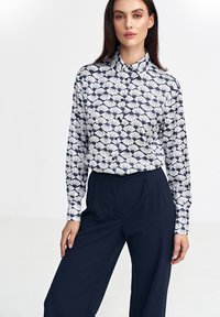 Koszula z długim rękawem w wzór, z granatowym i białym wzorem muszli, z kołnierzykiem i zapięciem na guziki, w połączeniu z granatowymi spodniami o szerokim kroju.