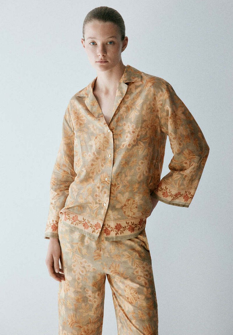 Femme portant une chemise boutonnée et un pantalon beige à motif floral assortis, debout avec une main dans la poche sur un fond uni.