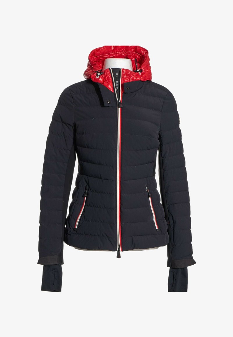 Veste matelassée noire avec capuche rouge, fermeture éclair avant avec bordure blanche et rouge, et poches latérales zippées sur un mannequin blanc.