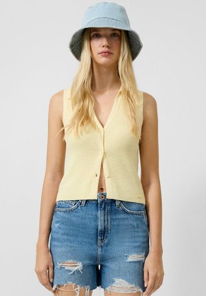 Jonge vrouw met een lichtgele mouwloze top met knopen, versleten denimshorts en een lichtblauwe bucket hat, staande tegen een effen achtergrond.