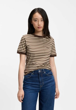 Jonge vrouw met lang donker haar die een bruin en beige gestreept T-shirt en blauwe spijkerbroek draagt, staand tegen een effen witte achtergrond.