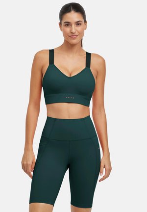 Femme portant un soutien-gorge de sport vert foncé et un short de cycliste taille haute assorti, debout sur un fond blanc uni.