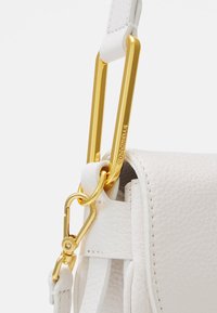 Valkoinen nahkalaukku, jossa on teksturoitu pinta, kultaiset metalliosat ja näyttävä lukko. Laukussa on leveä hihnakiinnike ja yksityiskohtainen ompele.