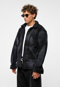 Zwarte sherpa-jacket met een knoopsluiting aan de voorkant, twee borstzakken en een kraag. Gedragen over een wit shirt, gecombineerd met zwarte broek.