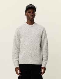 Pull en maille gris clair avec un motif texturé, encolure ronde et poignets côtelés. Porté avec un pantalon en velours côtelé noir et une casquette noire à motifs.