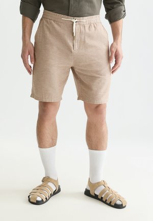 Mann trägt beige Shorts mit Kordelzug, weiße Sportsocken und beige Riemchensandalen, steht vor einem schlichten, hellen Hintergrund.