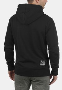 Schwarzer Kapuzenpullover mit einer weichen Textur, der ein Logo im unteren Rückenbereich sowie gerippte Bündchen und Saum aufweist.