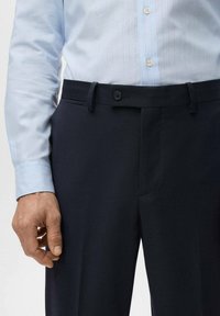 Granatowe eleganckie spodnie o gładkim kroju z przodu, zapinane na guziki, z szlufkami na pasek, zestawione z jasnoniebieską koszulą w paski.