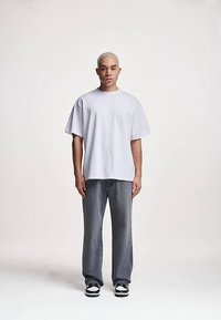 Volné bílé bavlněné tričko v kombinaci se šedými džínami s rovnými nohavicemi a černo-bílými teniskami, modelované osobou s krátkými vlasy.