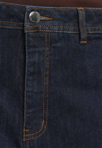 Detailní záběr na tmavě modré džínové kalhoty s oranžovým prošíváním, poutkem na opasek a kovovým knoflíkem s raženým textem.