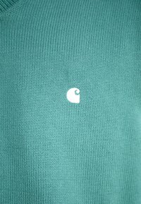 Pulover tricotat turcoaz cu un mic logo alb pe partea stângă. Textura este netedă, cu un model de tricotare strâns.