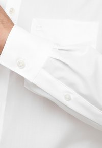 Witte overhemd met een ronde manchette en knoopsluiting, evenals een zak. De stof is soepel en egaal zonder zichtbare patronen.