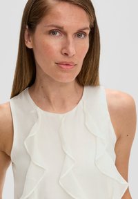 Weißes ärmelloses Blusenshirt mit Rüschendetails am Ausschnitt. Der Stoff wirkt leicht und glatt, mit einer sanften Textur.