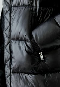 Zwarte pufferjas met een gequilt ontwerp, glanzende textuur en een ritszak met een zilverkleurige trekker aan de voorkant.