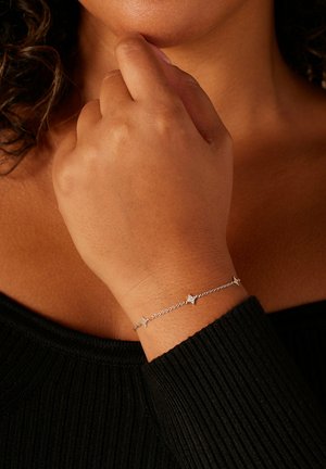 Zilveren armband met schakels van een ketting, met stervormige accenten. Draagt aan een pols, afstekend tegen een geribd zwart topje.