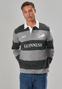 Gray and black striped rugby shirt with white collar, featuring "GUINNESS" logo and bird graphic. Model wears blue jeans.