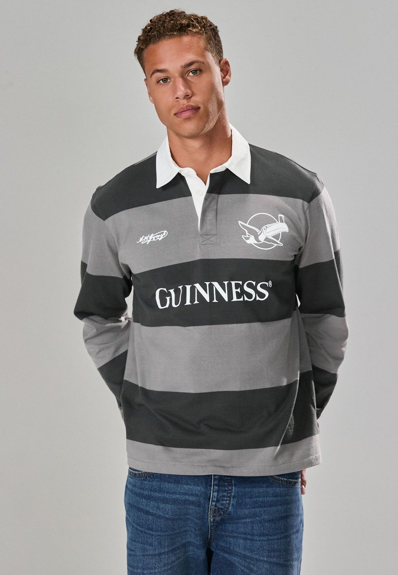 Gray and black striped rugby shirt with white collar, featuring "GUINNESS" logo and bird graphic. Model wears blue jeans.