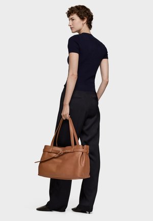 Brązowa skórzana torebka z dwoma długimi uchwytami, gładką fakturą i subtelnym detalem z klamrą. Modelka ubrana w czarne spodnie i krótkawy rękaw.
