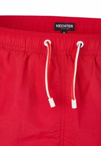 Shorts de bain rouges avec une taille élastique, dotés d'un cordon de serrage blanc et rouge. Fabriqués en tissu léger et texturé. Étiquette de marque visible.