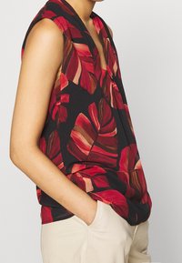 Ärmlös svart topp med stort tryck av röda och bruna tropiska blad, bärs med beige byxor, hand avslappnat i fickan, överkropp synlig.