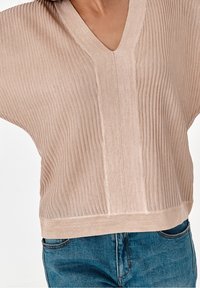 Haut en tricot côtelé beige clair avec un décolleté en V et des manches courtes, présentant un design texturé et une coupe décontractée, associé à un jean en denim bleu.