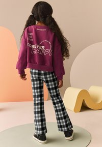 Fioletowa bluza z białymi wzorami tekstowymi, zestawiona z czarno-białymi spodniami w kratę typu dzwony i czarnymi sneakersami, na pastelowym tle.