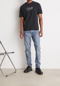 Svart bomullströja med en färgglad "Levi's"-logga, ihopparad med ljusblå denimjeans och svarta sneakers, mot en neutral bakgrund.