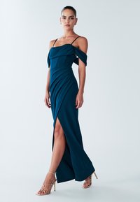 Teal avondjurk met off-shoulder mouwen, slim fit, gedrapeerde textuur en een hoge split aan de bovenkant van de dij, gecombineerd met gouden hoge hakken met bandjes.