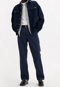 Veste en denim foncé portée sur une chemise grise à boutons, t-shirt blanc, pantalon bleu marine et chaussures habillées noires. Caractéristiques principales : poches, boutons et ceinture.