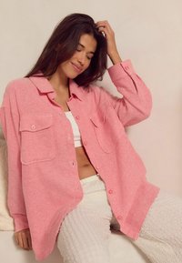 Lichtroze oversized overhemd met knopen en twee voorkeurszakken, vergezeld van een witte top en gestructureerde witte broek. Zachte stof.
