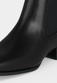 Bottine en cuir noir avec un talon bloc et un panneau latéral élastique, montrant une texture lisse et polie ainsi que des détails de couture.