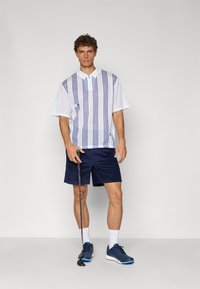 Hvit mesh polo skjorte med blå vertikale striper, kombinert med marineblå shorts. Modellen holder en golfklubbe, iført marineblå sneakers og hvite sokker.