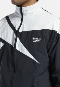 Man i svart och vitt Reebok zip-up jacka med hög krage och diagonala färgblockeringar på framsidan.