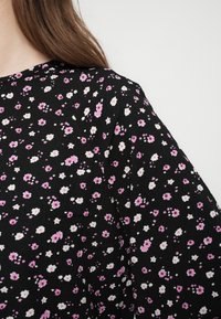 Donna che indossa un tessuto nero con piccolo motivo floreale rosa e bianco, che mostra la spalla e parte dei capelli su sfondo bianco.