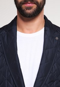 Marineblau gesteppte Jacke mit strukturiertem Kragen und silbernen Hardware-Elementen, getragen über einem schlichten weißen T-Shirt und präsentiert eine glatte Textur.