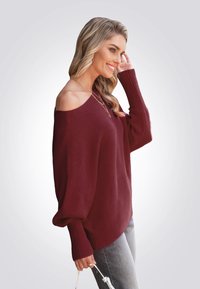 Kastanienrotes Off-Shoulder-Rippstrickoberteil mit langen Ärmeln, lässiger Passform, seitlichen Schlitzen und strukturiertem Stoff. Kombiniert mit hellen Jeans und Accessoires.