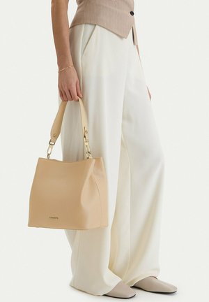 Person trägt weiße Hosen mit weitem Bein und beige Weste, hält eine beige Handtasche mit Metallbeschlägen am Griff.