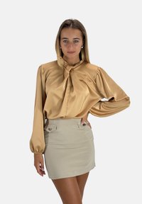 Blusa di raso beige con collo a cravatta e maniche a sbuffo, abbinata a una mini gonna aderente beige chiaro con bottoni decorativi.