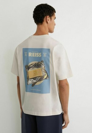 Muž v bielom tričku s potlačeným dizajnom masla na fólii a textom „REISS elite grade spread“ na chrbte, otočený chrbtom.