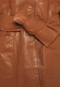 Brun läderjacka med ett brett bälte och en stor rosett, stora påsydda fickor och en slät, glänsande yta.