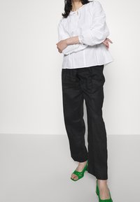 Blusa bianca a maniche lunghe con scollo rotondo e dettaglio con nodo, abbinata a pantaloni neri larghi e scarpe aperte verdi con design ritagliato.
