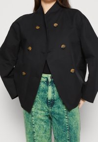 Zwarte dubbel-breasted blazer met goudkleurige knopen, een hoge kraag en een boxy silhouet. Draagt over groene acid-wash denim.