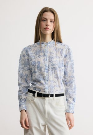 Fiatal nő kék-fehér mintás, gombos blúzban, dús vállrésszel, fehér nadrágot és fekete övet viselve, áll.