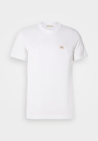 T-shirt bianco a maniche corte con collo rotondo e piccolo patch logo beige quadrato sul lato sinistro del petto.